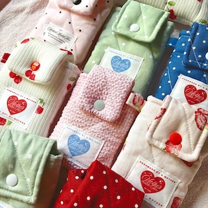 May include: A collection of handmade fabric pouches in various colors and patterns. Designs include polka dots, cherries, and strawberries. Each pouch has a flap closure with a button and a label that reads "Mother Made This."