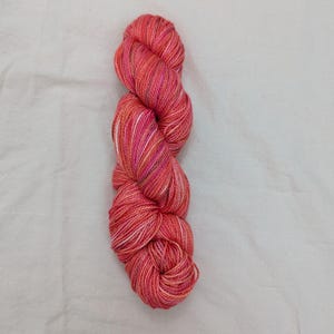 May include: A skein of hand-dyed yarn in shades of coral, pink, and orange. The yarn has subtle variations in color, with hints of white and yellow. The yarn is likely made of wool or a similar fiber, and is suitable for knitting or crocheting projects.
