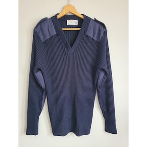 Peut inclure: Pull-over bleu marine à col en V avec une texture en tricot côtelé. Il présente des renforts bleu foncé sur les épaules et les coudes. Le vêtement est suspendu à un cintre en bois. Une petite étiquette est visible près de l'encolure.