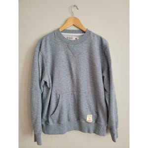 May include: A grey sweatshirt with a crew neck and a kangaroo pocket. The sweatshirt has long sleeves and a small patch on the lower right side. The garment is hanging on a wooden hanger against a neutral wall.