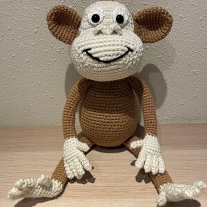 May include: A handmade crochet monkey doll with a smiling face. The monkey has a white head, hands, and feet, with brown ears, arms, legs, and body. The doll is sitting with its legs extended.