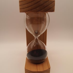 Puede incluir: Un reloj de arena en un marco de madera. La bombilla de vidrio transparente contiene arena negra. El marco de madera es de madera marrón claro.