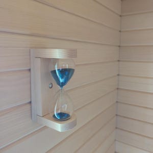 May include: An hourglass with blue liquid, mounted on a light wood wall. The hourglass is held by a wooden bracket. The background is made of light wood planks. The hourglass is a timekeeping device.