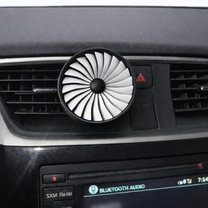 May include: A black and white car air vent diffuser with a turbine design. The diffuser is circular with white blades and a black center, mounted on a car's dashboard near the air vent.