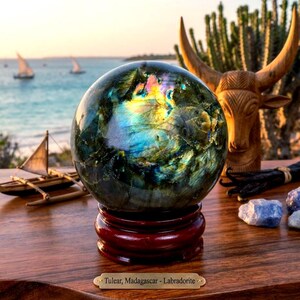 Puede incluir: Una esfera de labradorita pulida que muestra destellos iridiscentes azules, amarillos y verdes, sobre una base circular de madera oscura. Una pequeña placa dorada frente a la base presenta el texto: Tulear, Madagascar - Labradorite.