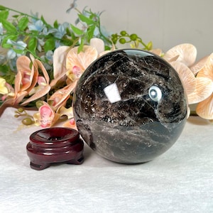 Könnte beinhalten: Eine große, polierte Rauchquarz-Kugel mit inneren Adern steht neben einem kleinen, dunkelroten Holzständer. Die Kugel reflektiert Licht, und der Hintergrund umfasst Blumenarrangements mit rosa und pfirsichfarbenen Blüten.