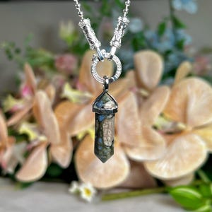 Könnte beinhalten: Eine silberne Halskette mit einem Anhänger aus blauem, grünem und braunem Kristall. Der Kristall hat die Form eines langen, spitzen Prismas. Die Halskette hat einen dekorativen Verschluss und eine zarte Kette. Der Hintergrund ist mit Blumen verschwommen.