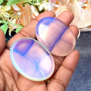 May include: Two oval-shaped, polished stones with a translucent appearance. The stones have a gradient of colours, including blue, purple, and pink. The stones are held in a hand, with flowers in the background. The stones are likely used for healing or meditation.
