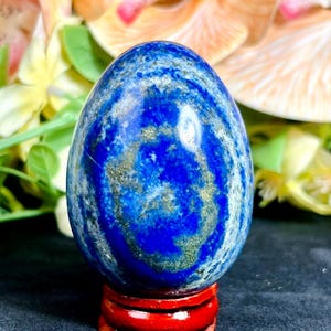 May include: A polished, egg-shaped lapis lazuli stone with swirling patterns of deep blue and gold. The gemstone is displayed on a small, red wooden stand, with a blurred floral background.
