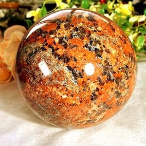 May include: A polished spherical stone featuring a vibrant mix of deep orange, burnt red, and dark charcoal grey mineral inclusions. The surface is smooth and reflective, showcasing a complex, crystalline texture with natural colour variations.