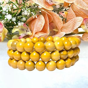 Op de afbeelding: Drie gestapelde armbanden gemaakt van geelgouden, ronde kralen. De kralen hebben een gepolijste, glanzende afwerking. De armbanden staan voor een vage achtergrond van perzikroze orchideeën en witte bloemen.