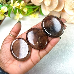Pode incluir: Três pedras de quartzo fumê polidas, em forma oval. As pedras são de cor marrom translúcido com padrões internos sutis. As pedras são seguradas em uma mão, com um anel de ouro visível em um dedo. O fundo inclui flores.