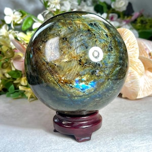 Puede incluir: Una esfera pulida de labradorita, que muestra destellos iridiscentes de azul, verde y dorado. La gema está montada sobre un soporte de madera rojo oscuro. La superficie de la esfera tiene patrones intrincados y una calidad reflectante.