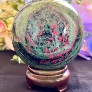 Puede incluir: Una piedra esférica pulida de rubí en zoisita con un patrón moteado de verde bosque profundo, magenta rosado vibrante e inclusiones negras. El orbe liso y reflectante se muestra descansando sobre un pequeño soporte circular de madera marrón.