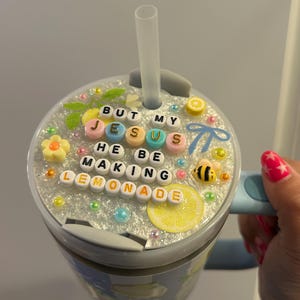 K&ouml;nnte beinhalten: Ein hellblauer Becher mit einem Deckel, der mit Perlen verziert ist, die "BUT MY JESUS HE BE MAKING LEMONADE" buchstabieren. Der Deckel ist mit einer Zitronenscheibe, einer Biene und anderen dekorativen Elementen verziert. Ein klarer Strohhalm ist in den Deckel eingesetzt.
