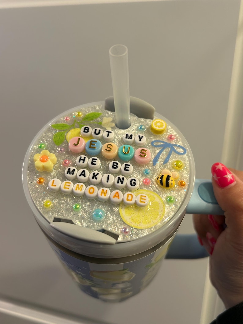 K&ouml;nnte beinhalten: Ein hellblauer Becher mit einem Deckel, der mit Perlen verziert ist, die "BUT MY JESUS HE BE MAKING LEMONADE" buchstabieren. Der Deckel ist mit einer Zitronenscheibe, einer Biene und anderen dekorativen Elementen verziert. Ein klarer Strohhalm ist in den Deckel eingesetzt.