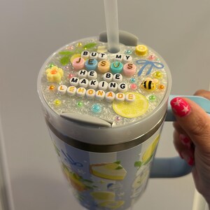 K&ouml;nnte beinhalten: Ein Edelstahlbecher mit hellblauem Griff und Strohhalm. Der Deckel ist mit Perlen verziert, die "BUT MY JESUS HE BE MAKING LEMONADE" buchstabieren. Der Becher hat ein Zitronen- und Kuchen-Design.