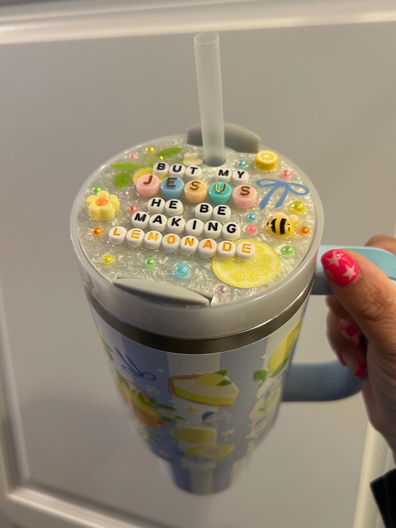 K&ouml;nnte beinhalten: Ein Edelstahlbecher mit hellblauem Griff und Strohhalm. Der Deckel ist mit Perlen verziert, die "BUT MY JESUS HE BE MAKING LEMONADE" buchstabieren. Der Becher hat ein Zitronen- und Kuchen-Design.