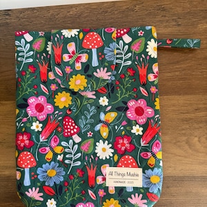 May include: A quilted fabric pouch featuring a dark green background with a colourful pattern of red mushrooms, pink and yellow flowers, and butterflies. A rectangular white label on the front reads All Things Mushie HANDMADE 2025.