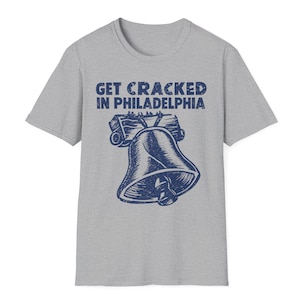 Op de afbeelding: Een lichtgrijs t-shirt met korte mouwen en een donkerblauwe afbeelding van de Liberty Bell met een houten juk. De tekst "GET CRACKED IN PHILADELPHIA" is in een verweerd donkerblauw lettertype boven de belafbeelding gedrukt.