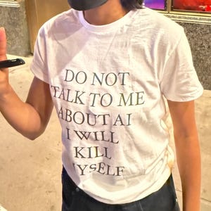 May include: A white short-sleeved crew neck t-shirt featuring black text printed on the front that reads: DO NOT TALK TO ME ABOUT AI I WILL KILL MYSELF. The shirt is made of a plain fabric and has a standard fit with a simple round neckline.