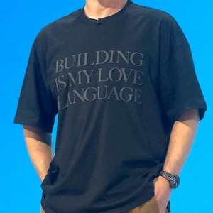 May include: A black short-sleeved t-shirt featuring the text "BUILDING IS MY LOVE LANGUAGE" printed in a dark, tonal font across the chest. The shirt has a crew neckline and a relaxed fit, with the text clearly visible against the black fabric.
