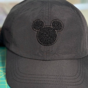 May include: A black baseball cap featuring a centred, embroidered black Mickey Mouse head silhouette. The cap has a structured crown, a curved brim with visible stitching, and a top button. A green cutting mat with grid lines is partially visible.