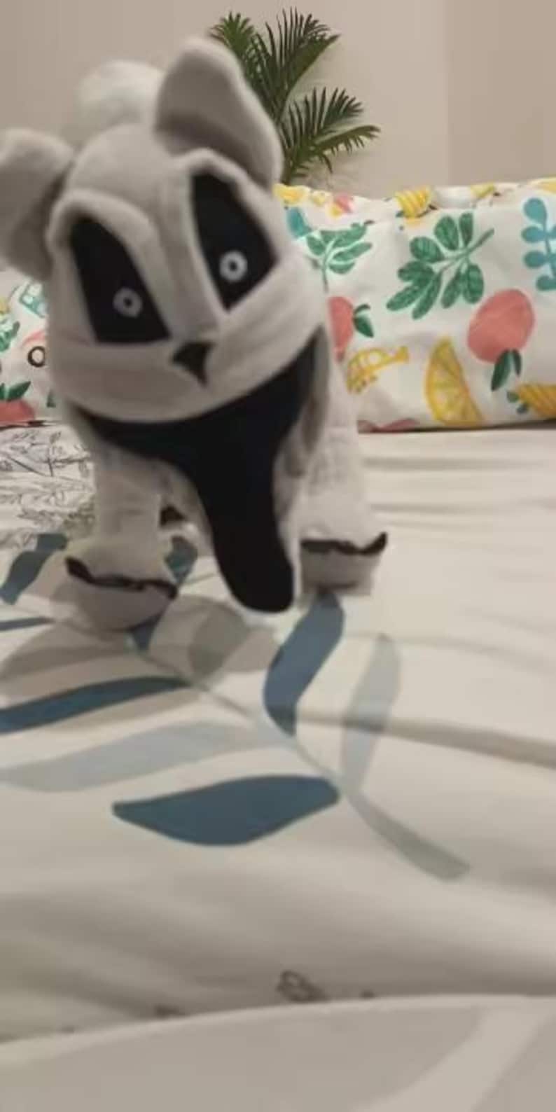 May include: A plush, gray and white toy animal with large black eyes and a black snout. The toy is posed on a bed with a floral patterned comforter. The toy's design resembles a cartoon character.