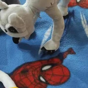 May include: A white plush toy in a quadrupedal stance, with black accents on its face and paws. The toy is on a blue blanket with a red and blue Spider-Man design. A yellow object is visible in the lower left corner.