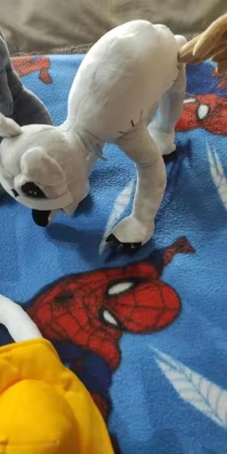 May include: A white plush toy in a quadrupedal stance, with black accents on its face and paws. The toy is on a blue blanket with a red and blue Spider-Man design. A yellow object is visible in the lower left corner.