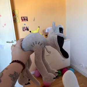 May include: A light gray plush toy with a black mouth and eyes, and long, thin legs. The toy is being held up in front of a light orange wall. Several colorful balloons are scattered on the floor.