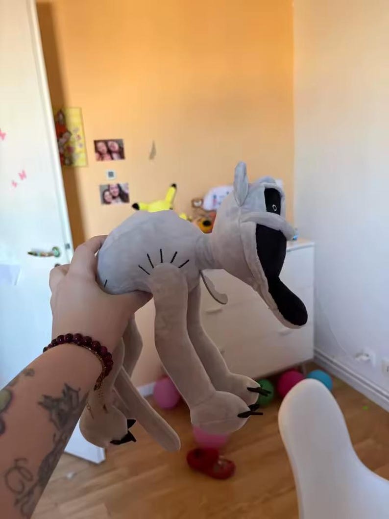 May include: A light gray plush toy with a black mouth and eyes, and long, thin legs. The toy is being held up in front of a light orange wall. Several colorful balloons are scattered on the floor.