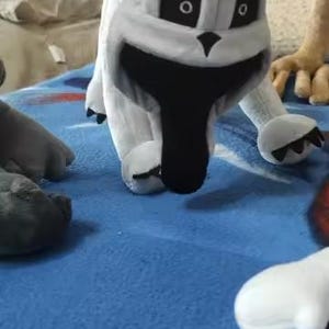 May include: A plush toy in the shape of a cartoon dog with a white body, black eyes, and a large black mouth. The dog is on a blue surface, with other plush toy parts visible.
