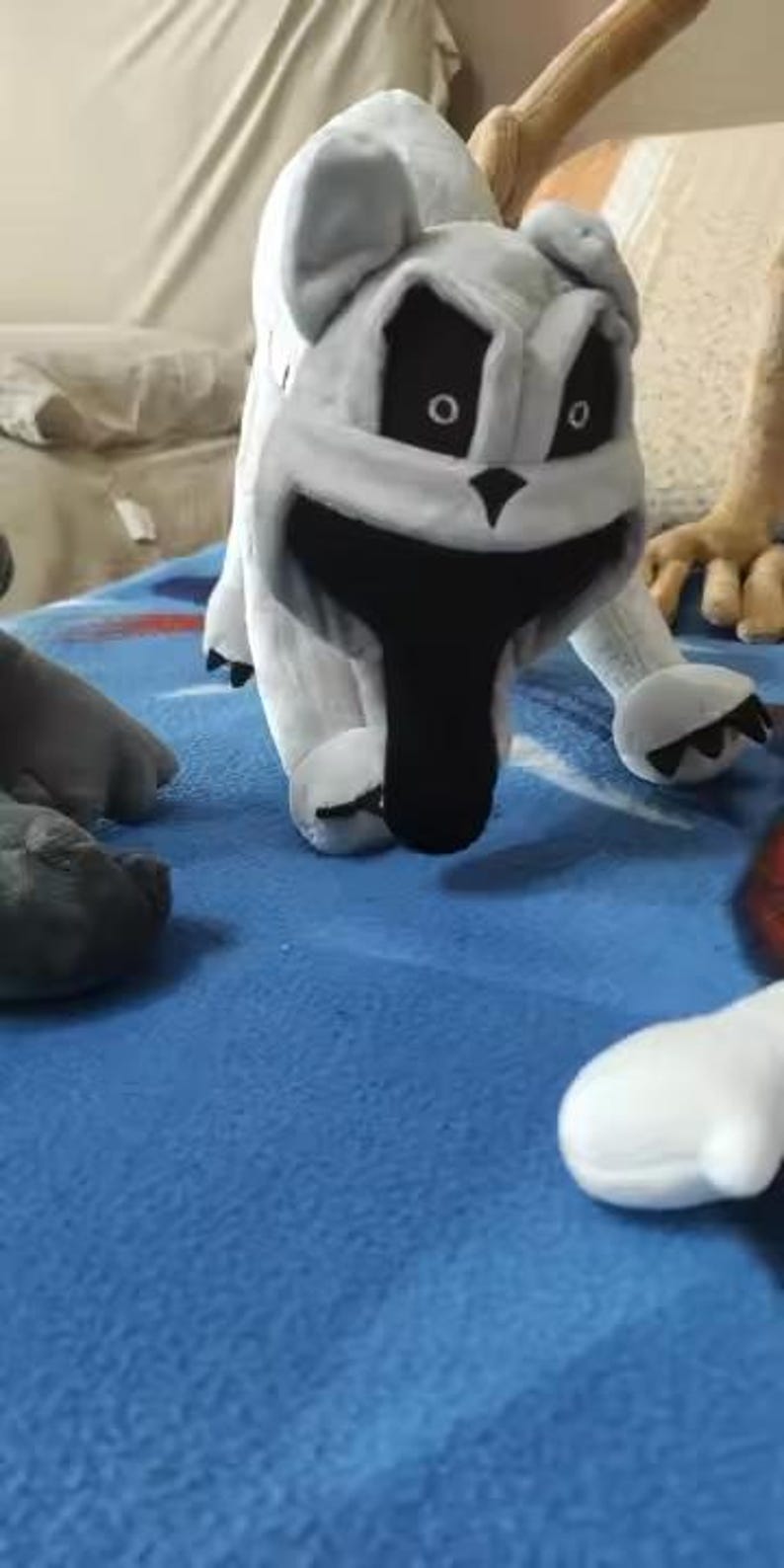 May include: A plush toy in the shape of a cartoon dog with a white body, black eyes, and a large black mouth. The dog is on a blue surface, with other plush toy parts visible.