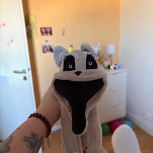 May include: A plush toy dog with a large, open mouth and black interior. The dog is primarily light gray with black eyes and a black nose. The toy has large ears and four paws. The background is a room with a yellow wall.