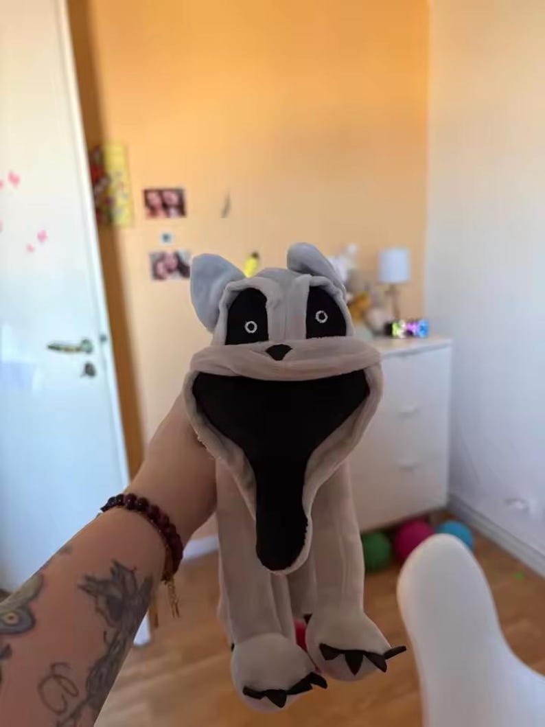 May include: A plush toy dog with a large, open mouth and black interior. The dog is primarily light gray with black eyes and a black nose. The toy has large ears and four paws. The background is a room with a yellow wall.