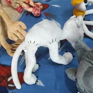 May include: A collection of plush toys, including a white cat with black markings, a yellow-hatted figure, and a gray shark. The toys are arranged on a blue patterned blanket, with other toy parts visible in the background.
