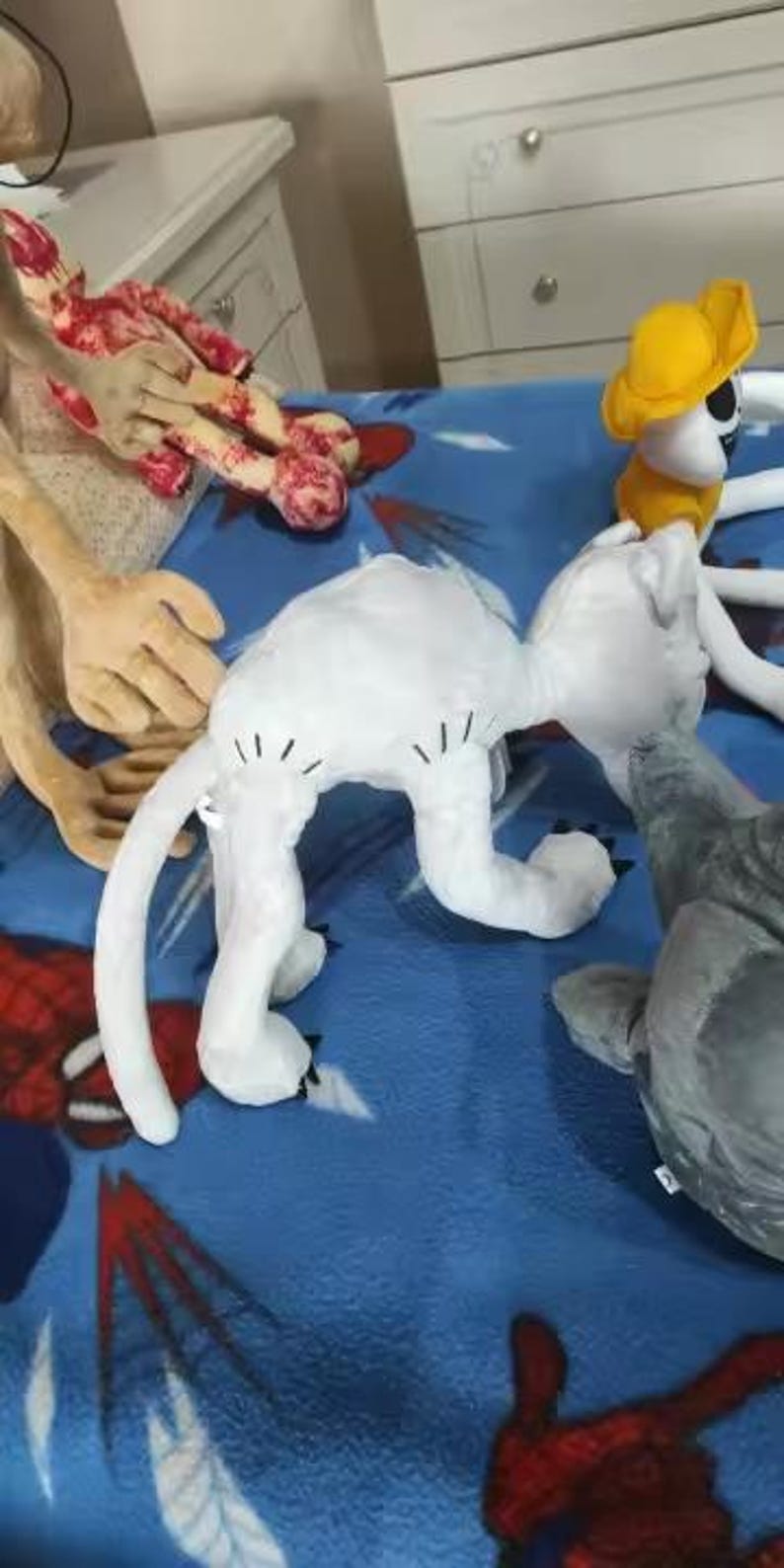 May include: A collection of plush toys, including a white cat with black markings, a yellow-hatted figure, and a gray shark. The toys are arranged on a blue patterned blanket, with other toy parts visible in the background.
