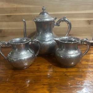 May include: A silver-toned metal tea set consisting of a lidded teapot, a sugar bowl with two handles, and a creamer pitcher. Each piece features an engraved floral pattern, a scalloped rim, and ornate, curved handles with detailed scrollwork.