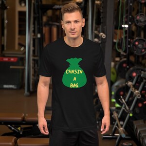 May include: Black t-shirt with a green money bag graphic and the text "CHASIN A BAG" in yellow. The shirt is made of a soft material and is suitable for casual wear.