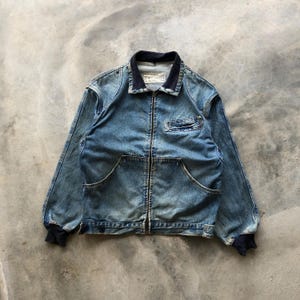 Könnte beinhalten: Eine blaue Jeansjacke mit durchgehendem Frontreißverschluss, einem dunkelblauen gerippten Kragen und passenden gerippten Bündchen. Die Jacke verfügt über zwei untere Vordertaschen und eine einzelne Reißverschlusstasche auf der linken Brust.