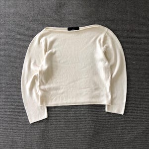 May include: A long-sleeved, off-white knit sweater featuring a wide boat neckline and a straight hem. The garment is constructed from a smooth, fine-gauge knit fabric and includes a small black interior brand label at the center of the neckline.