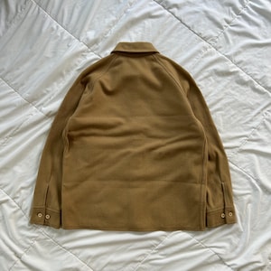 K&ouml;nnte beinhalten: Eine beigefarbene Langarmjacke mit klassischem Kragen, Raglan&auml;rmeln und gekn&ouml;pften Manschetten. Das Kleidungsst&uuml;ck besteht aus einem einfarbigen Stoff mit glatter Textur und zeigt eine klare, strukturierte R&uuml;ckansicht mit sichtbaren N&auml;hten.