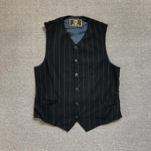 May include: A black vest featuring thin white pinstripes, a V-neckline, and a five-button front closure. The garment includes two lower welt pockets and a single chest pocket, with a dark grey interior lining and a branded label at the neck.