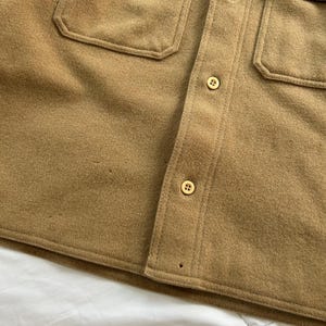 K&ouml;nnte beinhalten: Ein beiges Hemd mit Knopfleiste und weicher, gefilzter Textur, zwei Brusttaschen mit gen&auml;hten Klappen und einer zentralen Knopfleiste mit zwei sichtbaren hellen Vier-Loch-Kn&ouml;pfen. Das Kleidungsst&uuml;ck hat einen geraden Saum und sichtbare N&auml;hte.