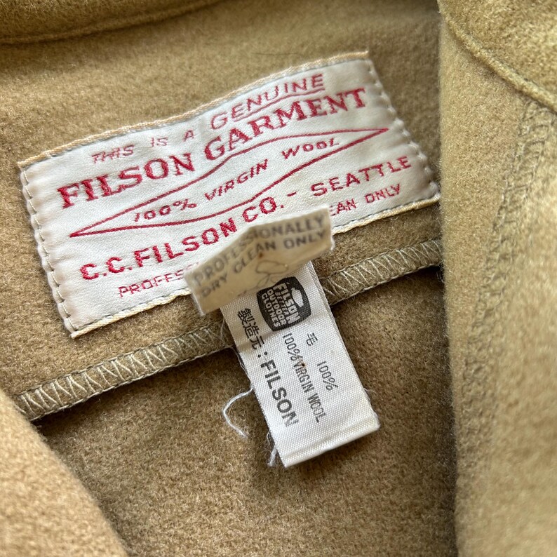 K&ouml;nnte beinhalten: Ein beigefarbenes Kleidungsst&uuml;ck aus Wolle mit einem wei&szlig;en rechteckigen Etikett mit rotem Text: THIS IS A GENUINE FILSON GARMENT 100% VIRGIN WOOL C.C. FILSON CO. SEATTLE. Ein kleineres wei&szlig;es Pflegeetikett darunter zeigt 100% VIRGIN WOOL und FILSON.