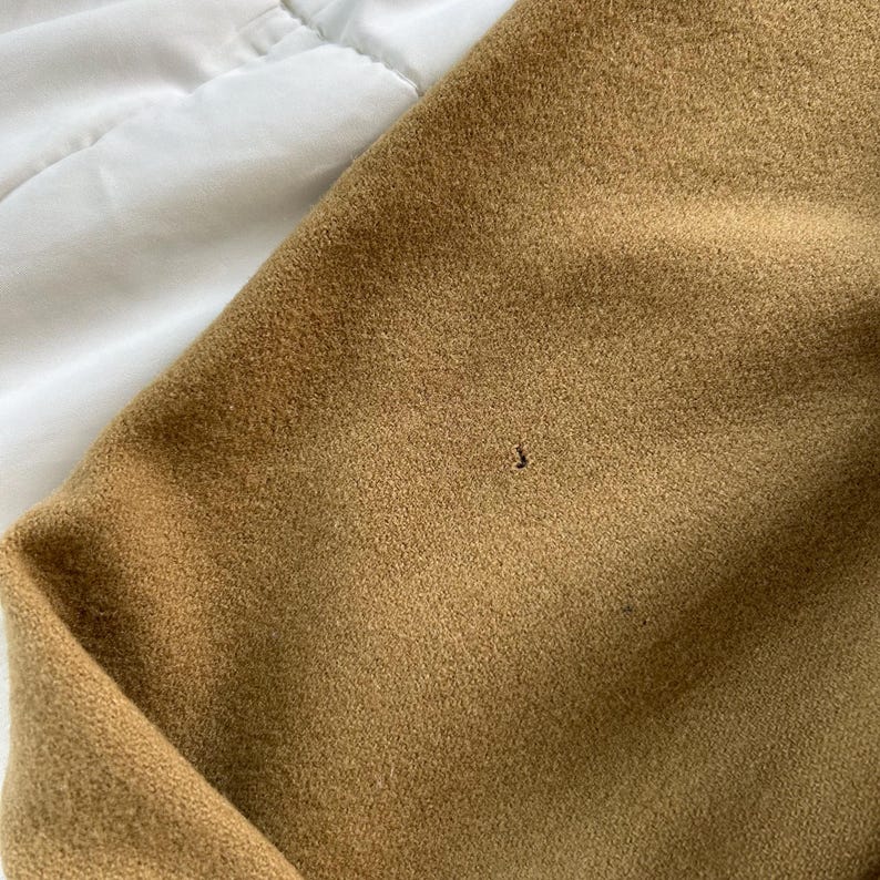 K&ouml;nnte beinhalten: Ein beigefarbener Stoff mit einer weichen, gefilzten Textur. Das Material weist ein kleines, einzelnes dunkles Loch in der Mitte der Oberfl&auml;che auf, das eine leichte Unvollkommenheit im Gewebe des Textils zeigt.