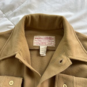 K&ouml;nnte beinhalten: Ein beigefarbenes Wollhemd mit spitzem Kragen, Knopfleiste und zwei Brusttaschen. Ein wei&szlig;es rechteckiges Etikett im Nackenbereich tr&auml;gt die Aufschrift: This is a Genuine Filson Garment, 100% Virgin Wool, C.C. Filson Co. Seattle.