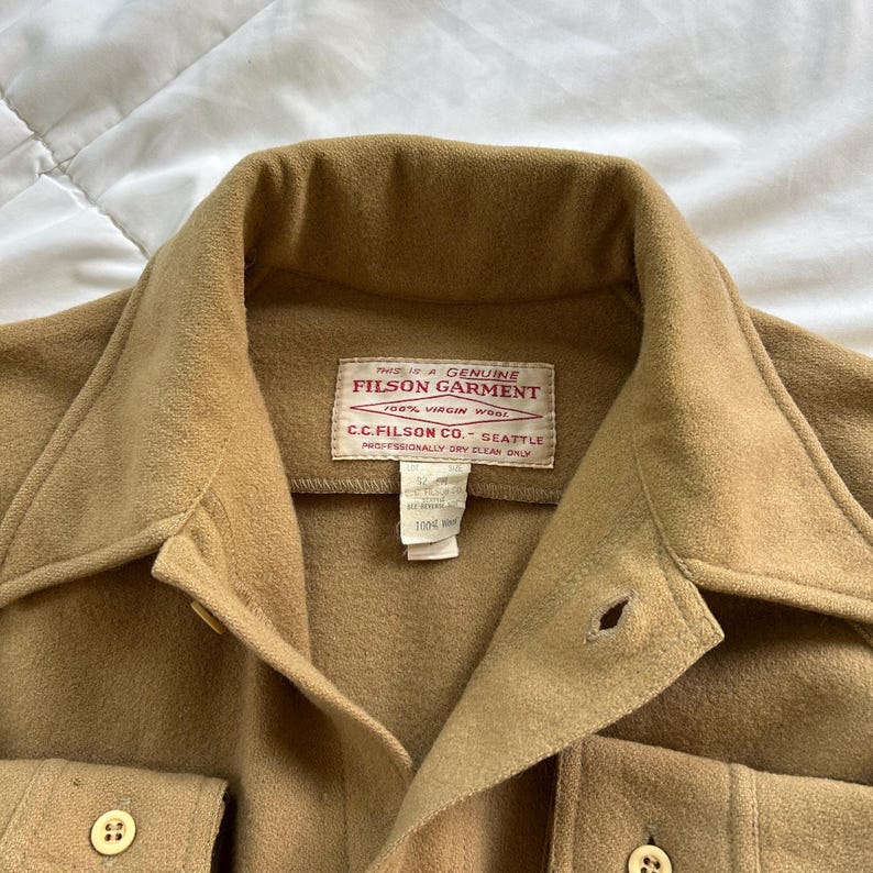 K&ouml;nnte beinhalten: Ein beigefarbenes Wollhemd mit spitzem Kragen, Knopfleiste und zwei Brusttaschen. Ein wei&szlig;es rechteckiges Etikett im Nackenbereich tr&auml;gt die Aufschrift: This is a Genuine Filson Garment, 100% Virgin Wool, C.C. Filson Co. Seattle.