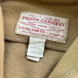 K&ouml;nnte beinhalten: Ein beigefarbenes Kleidungsst&uuml;ck aus Wolle mit einem wei&szlig;en rechteckigen Etikett mit rotem Text THIS IS A GENUINE FILSON GARMENT 100% VIRGIN WOOL C.C. FILSON CO. SEATTLE PROFESSIONALLY DRY CLEAN ONLY und einem kleineren Etikett f&uuml;r Gr&ouml;&szlig;e 32 SM und 100% Wool.